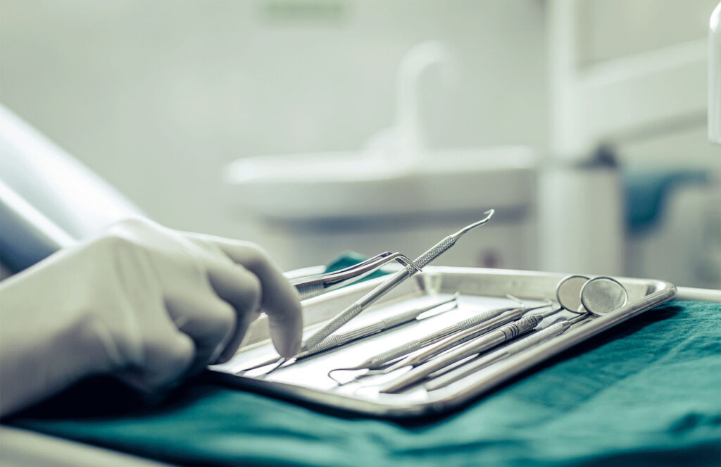 A gloved hand reaching for dental instruments arranged on a metal tray in a clinical setting, indicating preparation for a dental procedure.